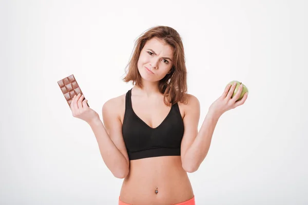 Confuso jovem fitness senhora segurando maçã e chocolate — Fotografia de Stock