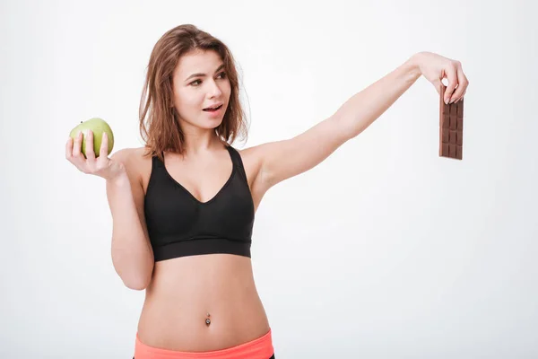 Sonriente joven fitness mujer sosteniendo manzana y chocolate — Foto de Stock