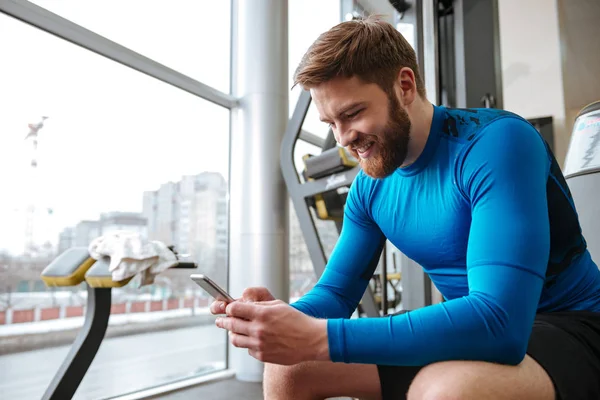 Fröhlicher junger Sportler sitzt im Fitnessstudio und schaut aufs Handy. — Stockfoto