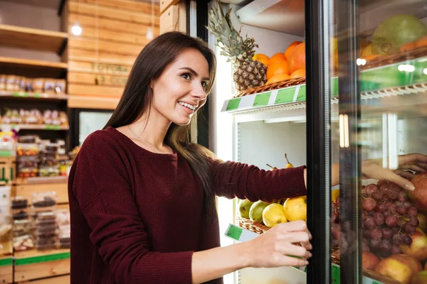 Улыбающаяся женщина выбирает и покупает фрукты в продуктовом магазине — стоковое фото