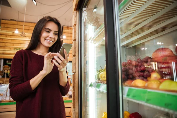 Женщина пользуется мобильным телефоном и покупает фрукты в продуктовом магазине — стоковое фото