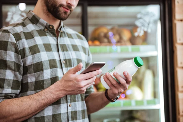 Bakkal dükkanında smartphone kullanarak süt şişesi olan adam — Stok fotoğraf