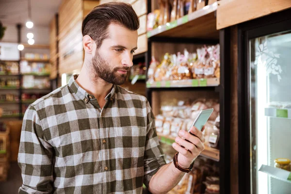 Guapo barbudo joven usando el teléfono celular en la tienda de comestibles —  Fotos de Stock