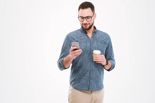 Cheerful bearded man standing with cup of coffee and smartphone — Stock Photo, Image