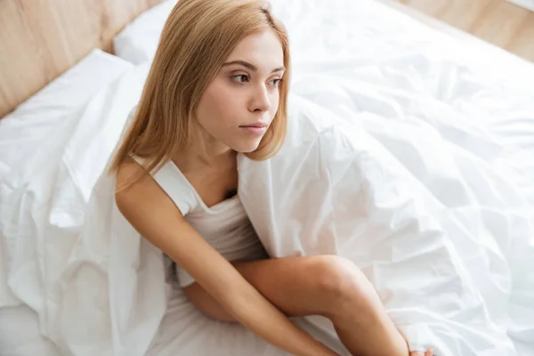 Mujer joven pensativa sentada y pensando en la cama en casa —  Fotos de Stock