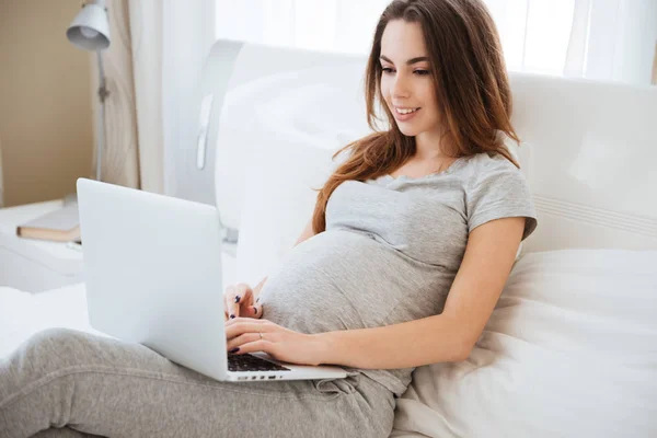 Joven embarazada alegre mintiendo y usando el ordenador portátil en la cama — Foto de Stock