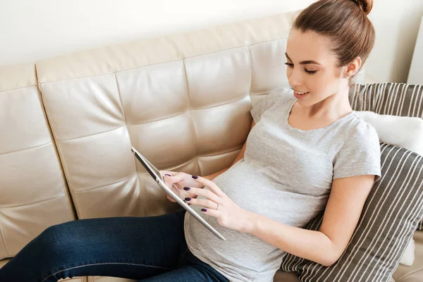 Happy pregnant young woman sitting on sofa and using tablet — Stock Photo, Image