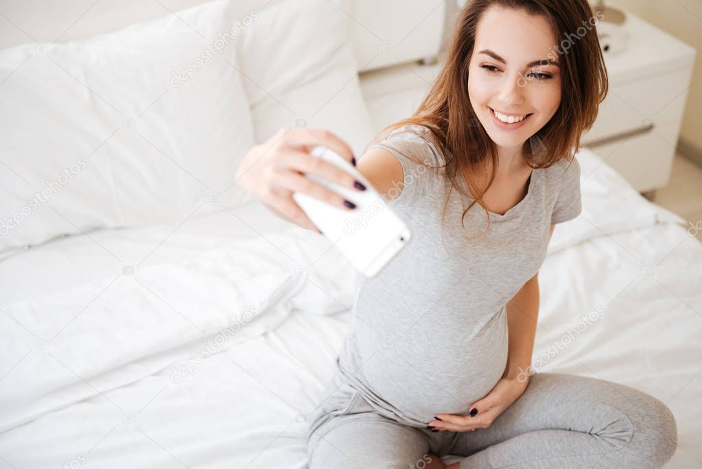 Cheerful pregnant woman sitting and taking selfie with mobile phone