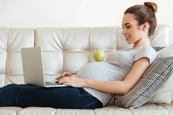 Joven embarazada alegre comiendo manzana y utilizando el ordenador portátil — Foto de Stock