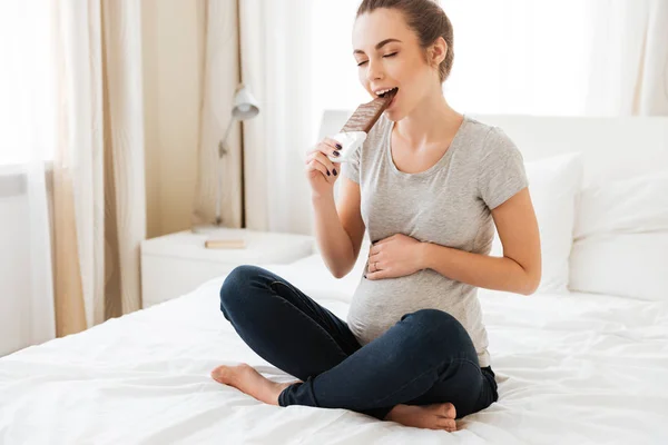 Feliz embarazada joven comiendo barra de chocolate en la cama —  Fotos de Stock