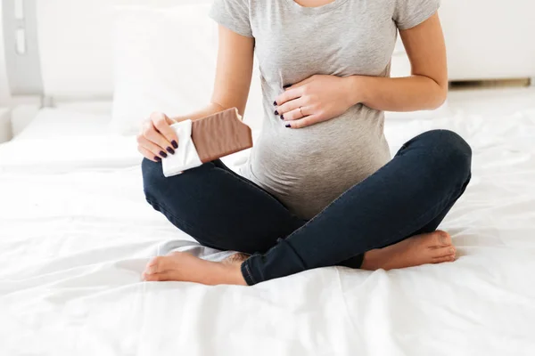 Pregnant young woman sitting and eating chocolate bar on bed — Stock Photo, Image