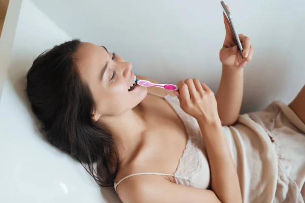 Веселая женщина вздымает зубы и использует мобильный телефон в ванной — стоковое фото