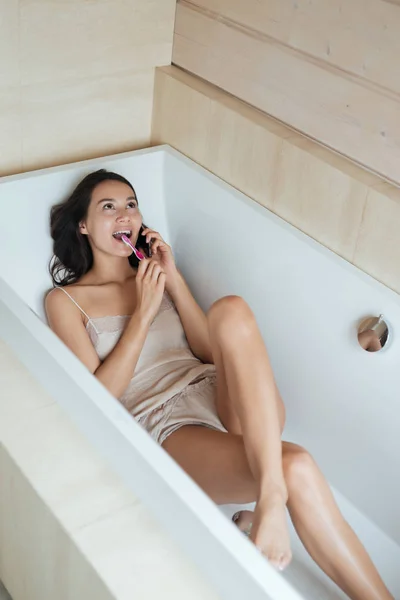 Mujer hablando por teléfono celular y cepillándose los dientes en la bañera — Foto de Stock