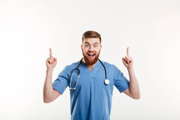 Excited young male doctor pointing two fingers up at copyspace — Stock Photo, Image