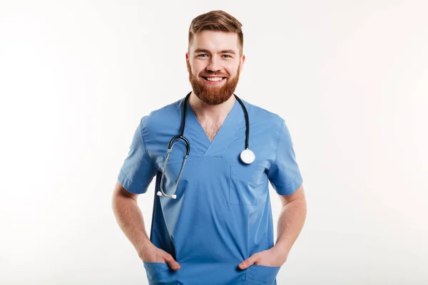 Médico varón sonriente de pie con estetoscopio y de la mano en los bolsillos —  Fotos de Stock