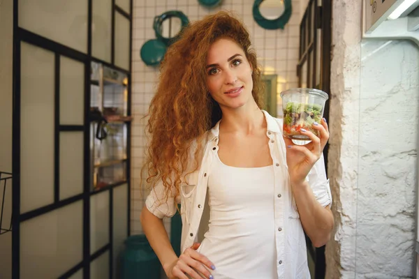 Sonriente pelirroja joven de pie en la cafetería mientras sostiene la ensalada . — Foto de Stock