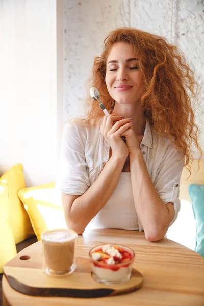 カフェでデザートを食べながら座っている赤毛の豪華な若い女性. — ストック写真