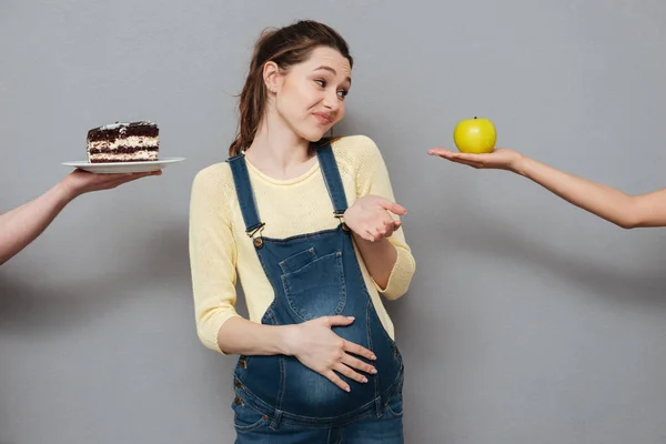 Junge verwirrte Schwangere hat die Wahl zwischen Apfel und Sahnetorte — Stockfoto