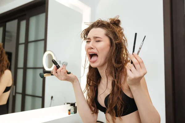 Грустная смущенная женщина с кисточками и косметикой плачет в ванной комнате — стоковое фото