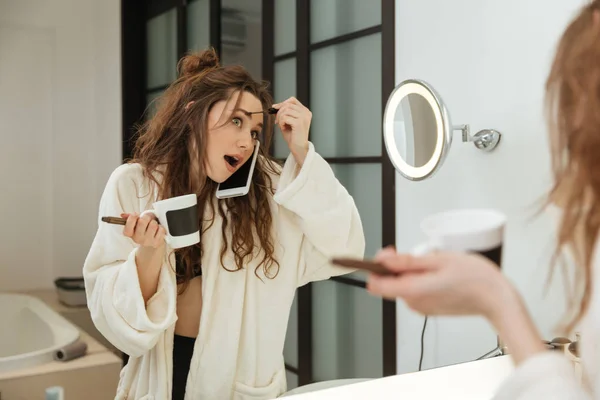 Женщина делает макияж и разговаривает по мобильному телефону в ванной комнате — стоковое фото