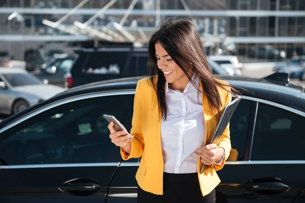 Joven empresaria alegre sosteniendo la carpeta y usando el teléfono celular al aire libre —  Fotos de Stock