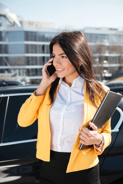 Feliz empresaria sosteniendo la carpeta y hablando en el teléfono móvil al aire libre —  Fotos de Stock