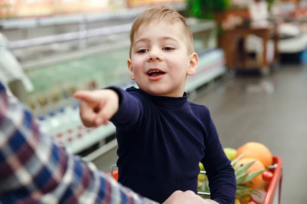 Happy boy sitting on shopping trolley and pointing away — Stock Photo, Image