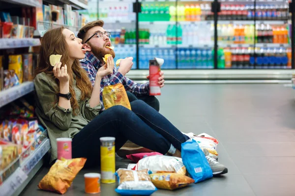 Pareja sentada en el piso del supermercado y comiendo bocadillos —  Fotos de Stock