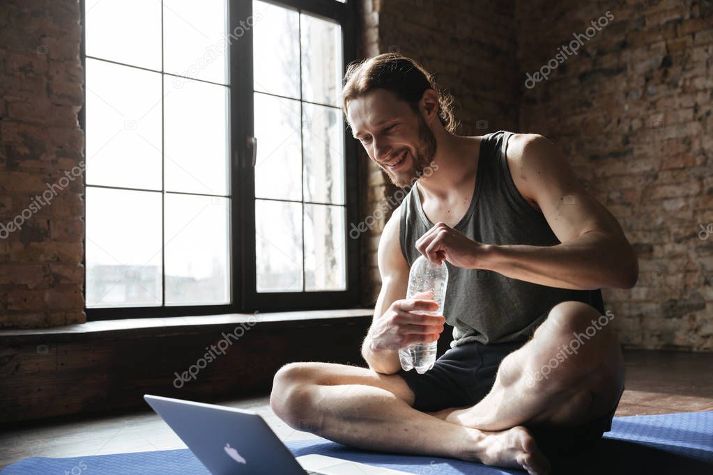 Happy strong sportsman drinking water on floor in gym