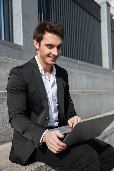 Smiling young businessman sitting outdoors using laptop — Stock Photo, Image