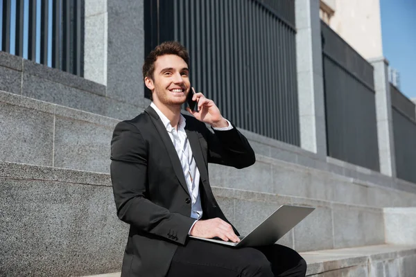 Hombre alegre sentado con el teléfono y el ordenador portátil — Foto de Stock