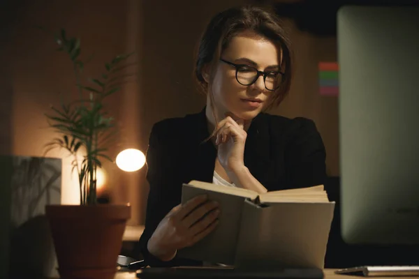 Serious young lady designer sitting indoors at night reading book. — Stock Photo, Image