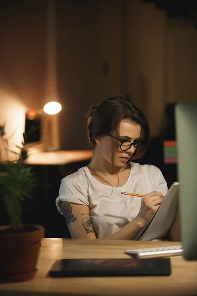 Concentrated young lady designer sitting indoors at night writing notes. — Stock Photo, Image