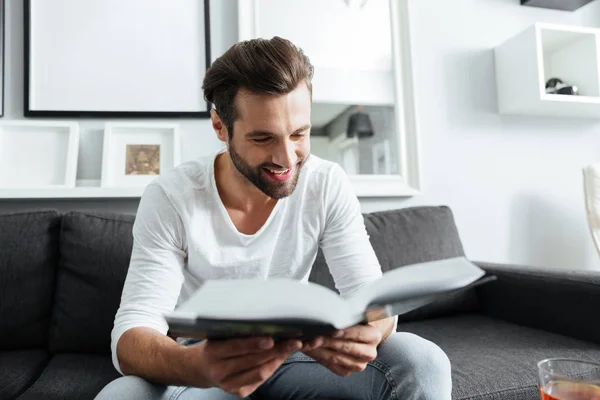 Fröhlicher Mann sitzt auf Sofa und liest Buch — Stockfoto