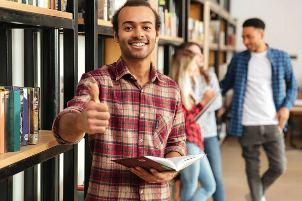 Jovem estudante feliz homem de pé na biblioteca mostrando polegares para cima . — Fotografia de Stock