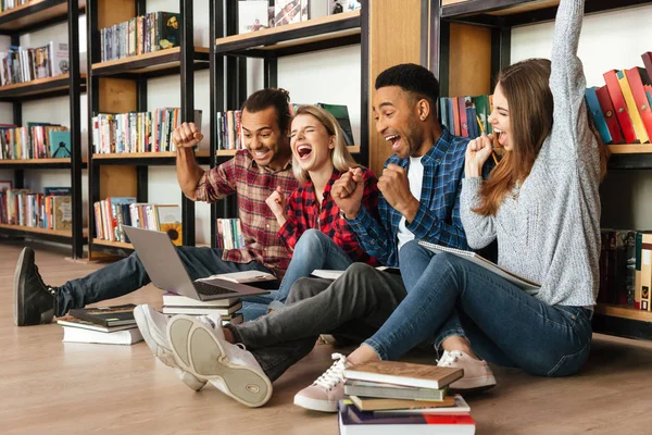 Allievi felici seduti in biblioteca sul pavimento utilizzando il computer portatile — Foto Stock