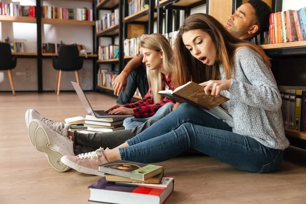 Estudiantes concentrados sentados en la biblioteca en el suelo usando portátil —  Fotos de Stock