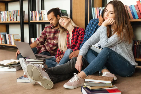 Estudantes cansados sentados na biblioteca no chão usando o computador portátil — Fotografia de Stock