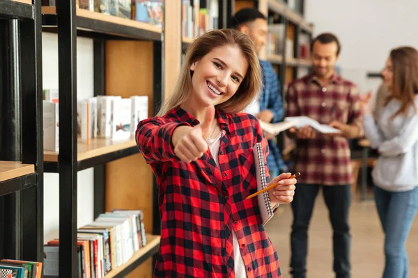 Jovem mulher sorridente estudante de pé na biblioteca mostrando polegares para cima — Fotografia de Stock