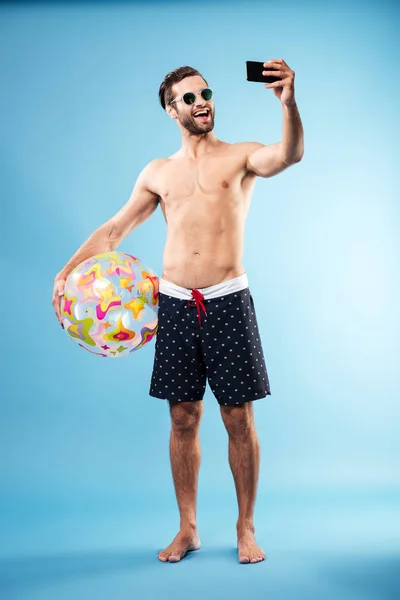 Feliz chico sin camisa sosteniendo pelota de playa y haciendo selfie — Foto de Stock