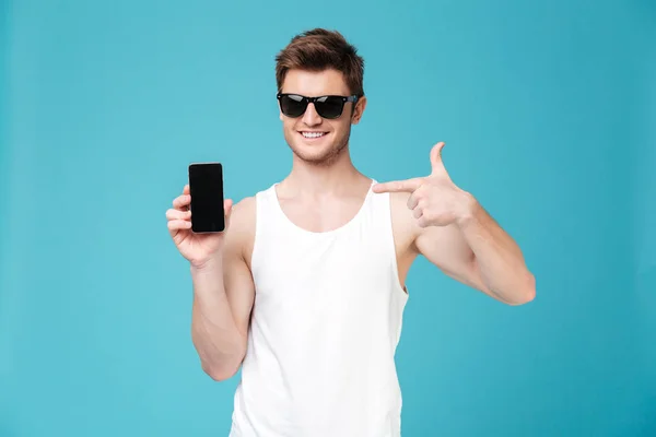 Hombre joven apuntando a la pantalla en blanco del teléfono inteligente aislado sobre azul — Foto de Stock