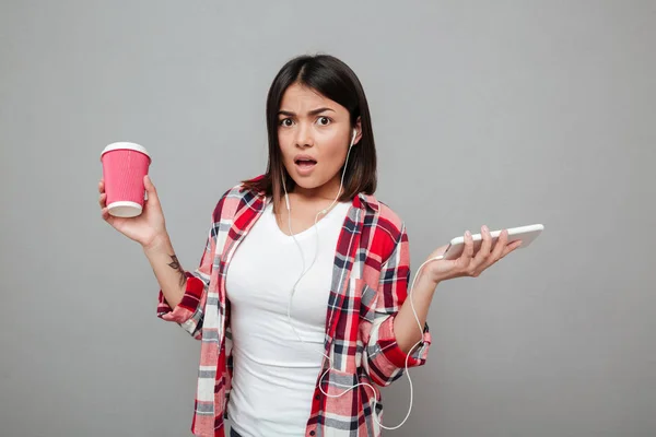 Mujer confusa sosteniendo una taza de café y escuchando música . — Foto de Stock