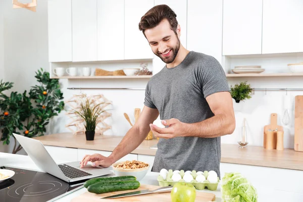 Homem com laptop preparando comida na cozinha — Fotografia de Stock
