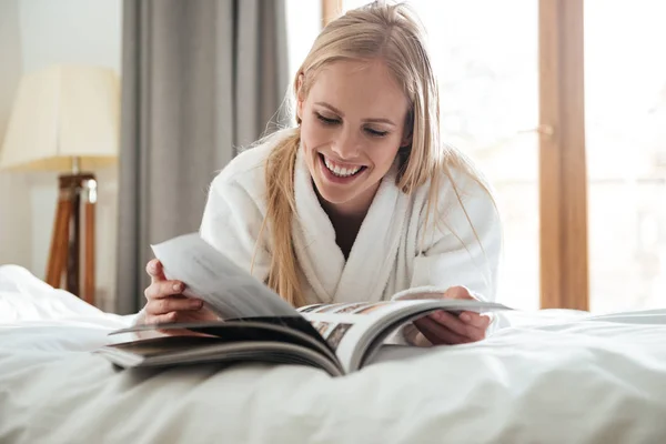 Junge blonde Frau liest Magazin, während sie auf dem Bett liegt — Stockfoto