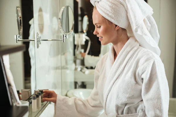 Mujer en albornoz mirando cosméticos cerca del espejo — Foto de Stock