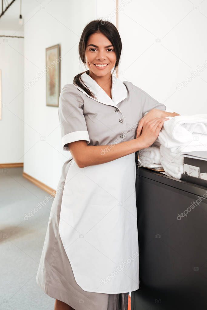 Smiling housekeeping worker standing with bedclothes linen in cart