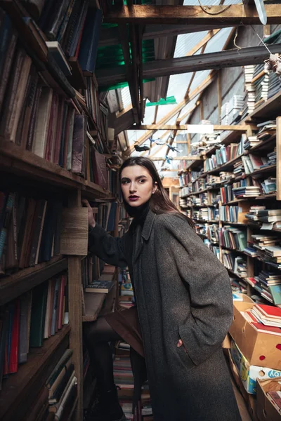 Mulher na moda subindo em uma escada em um armazém de livro — Fotografia de Stock