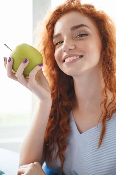 Wesoły redhead Pani siedzi przy stole w pomieszczeniu jedzenia apple — Zdjęcie stockowe