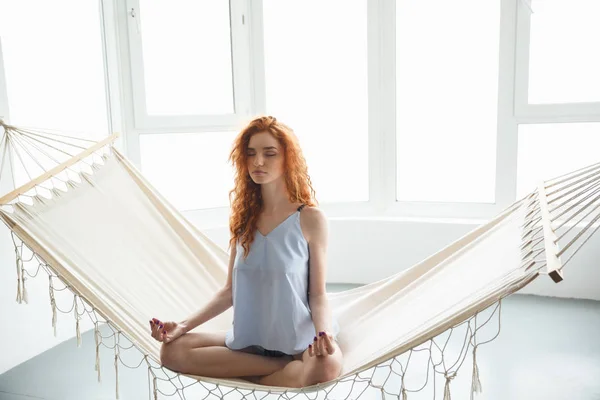 Geconcentreerde jonge roodharige dame zittend op de hangmat mediteren — Stockfoto