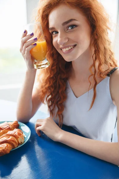 Sonriente pelirroja sentada en la mesa bebiendo jugo — Foto de Stock